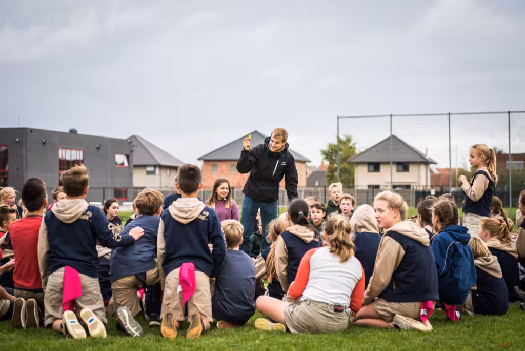 Groep kinderen luistert naar leraar op sportveld.